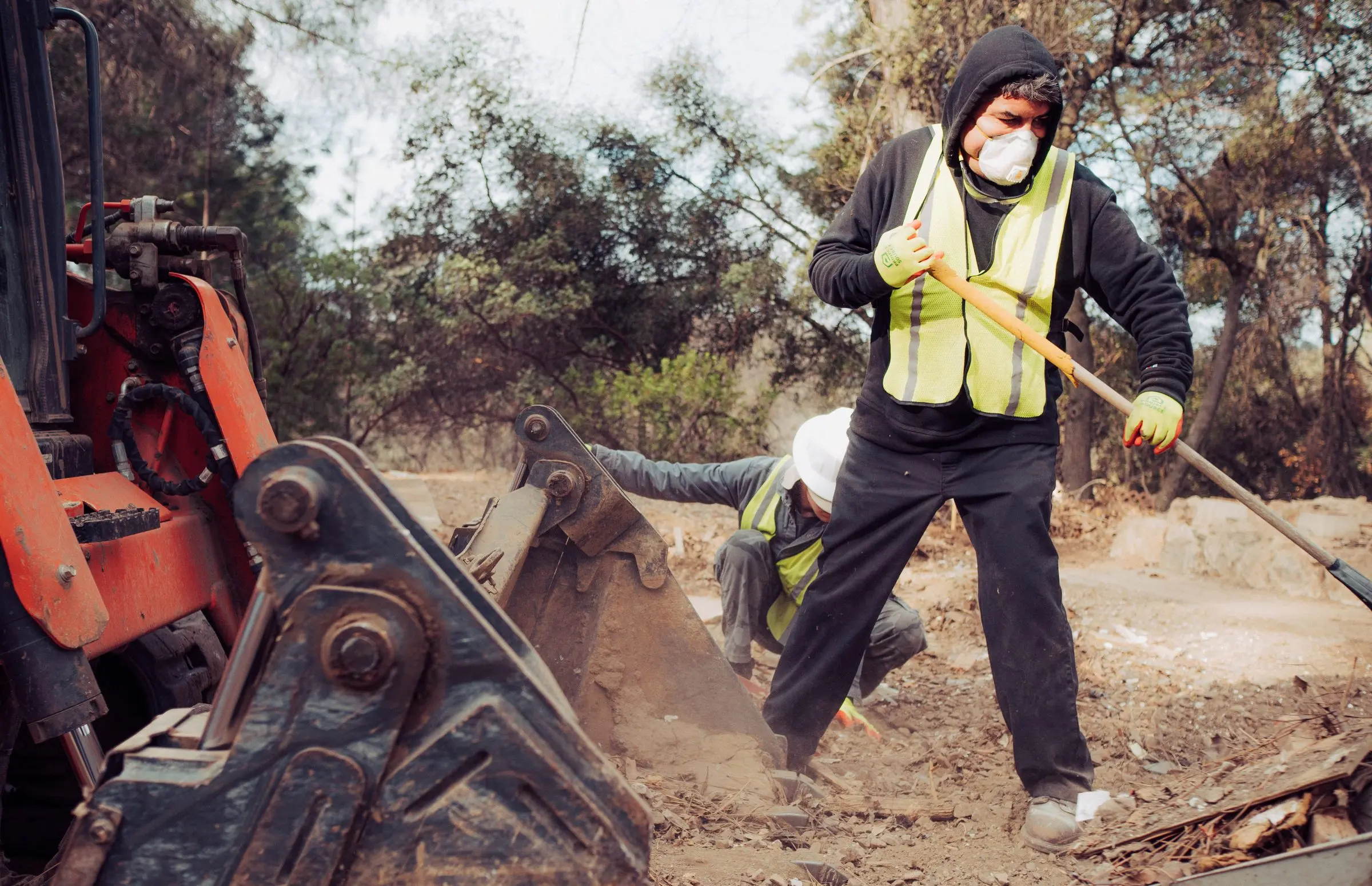 Crew member in a respirator shoveling debris next to a skid steer bucket