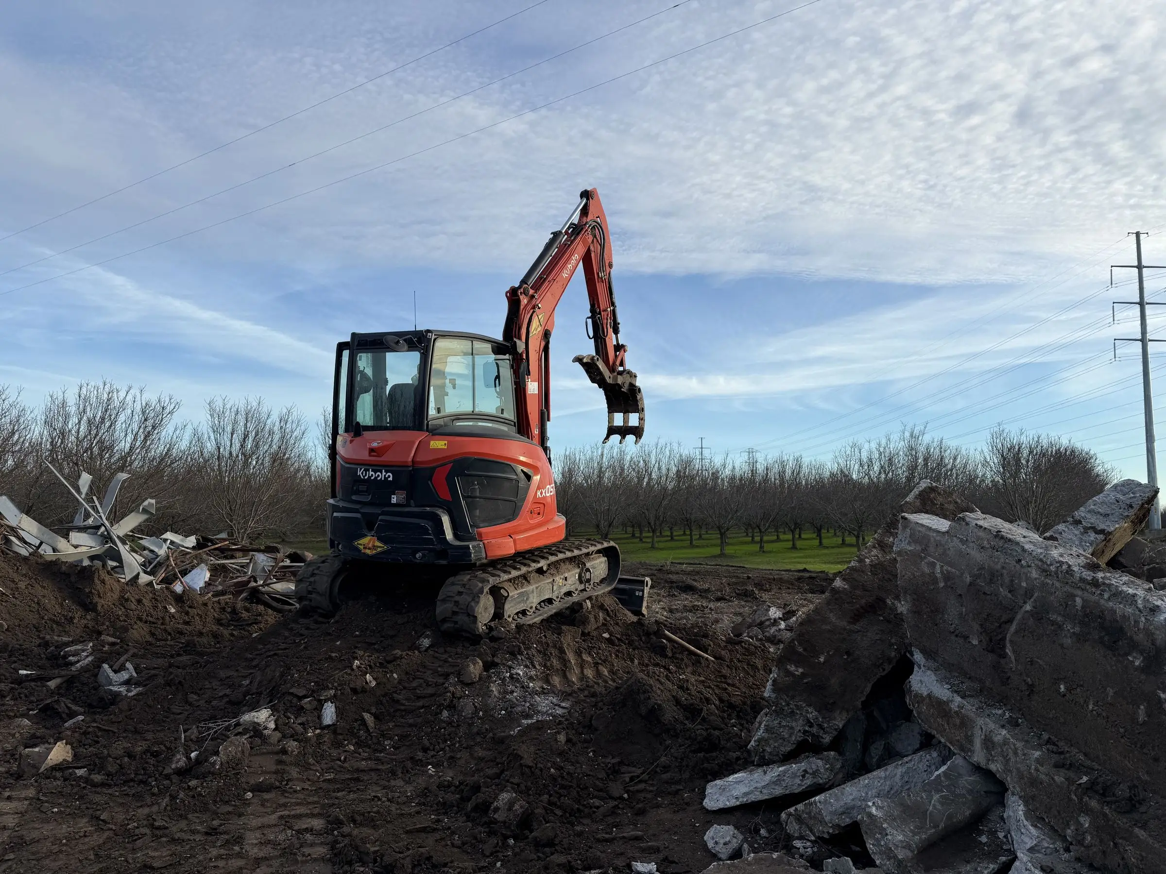 Kubota excavator beside broken concrete slabs on a rural lot