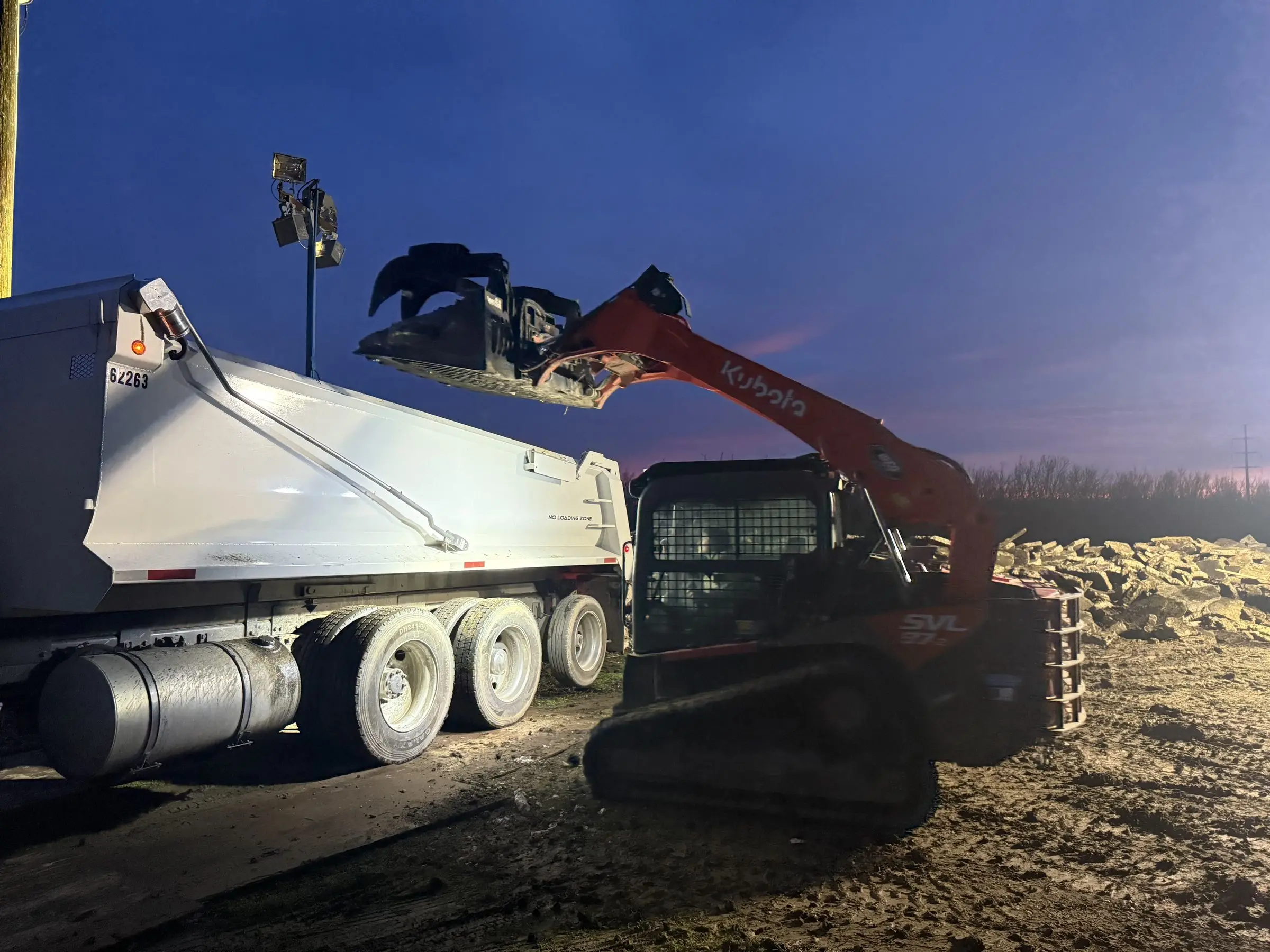 Skid steer loading a dump truck at dusk