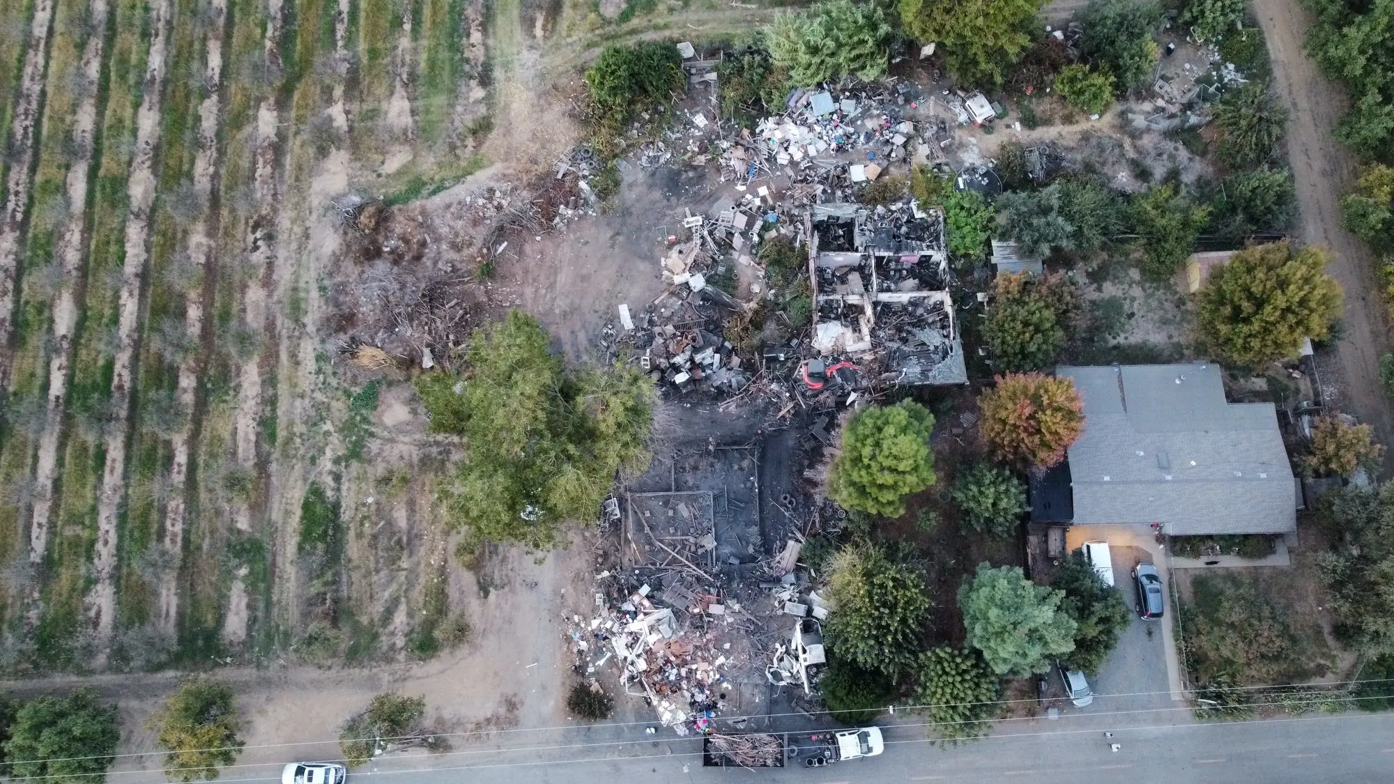 Aerial view of burned structures and debris beside an orchard and a neighboring house