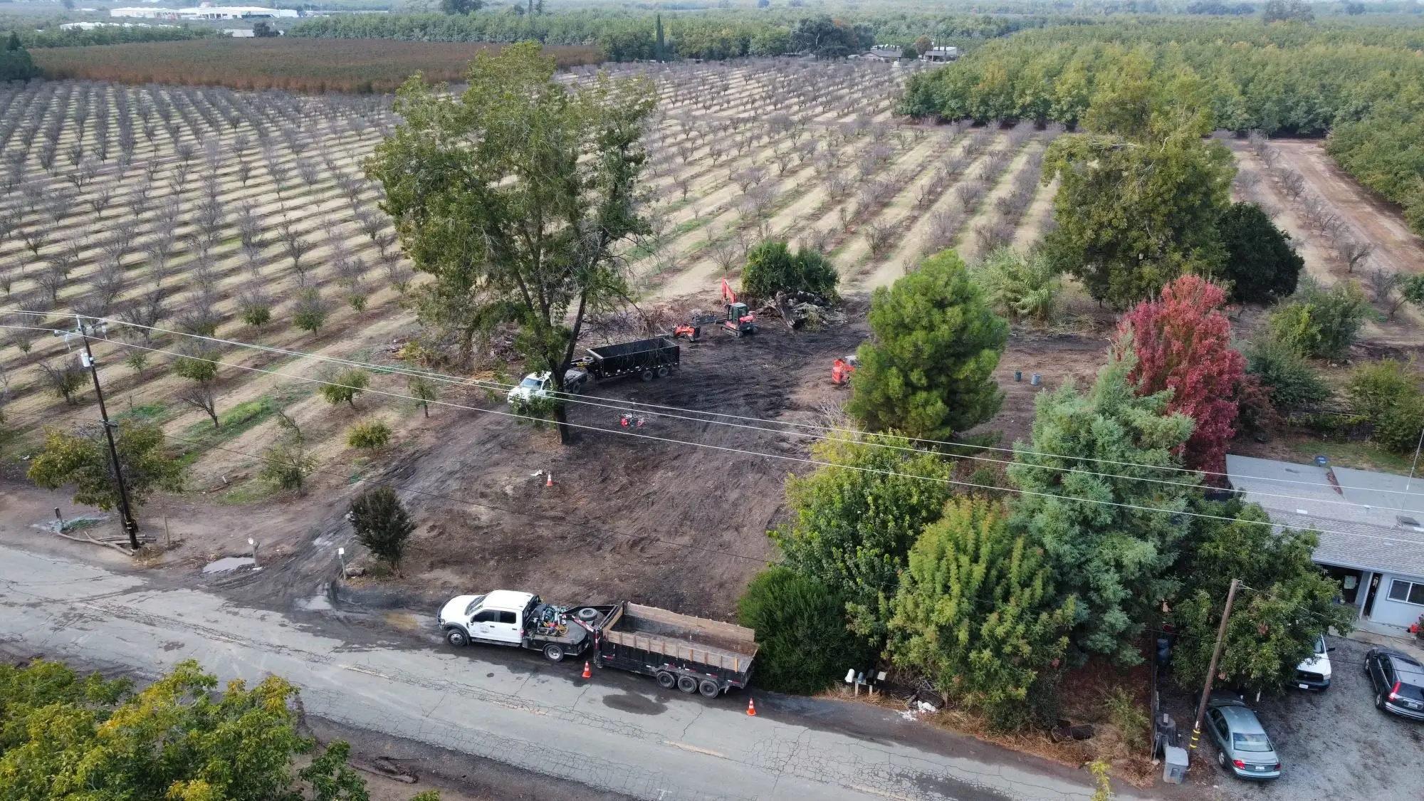 Aerial view of a cleared rural property beside an orchard with trucks parked on the road