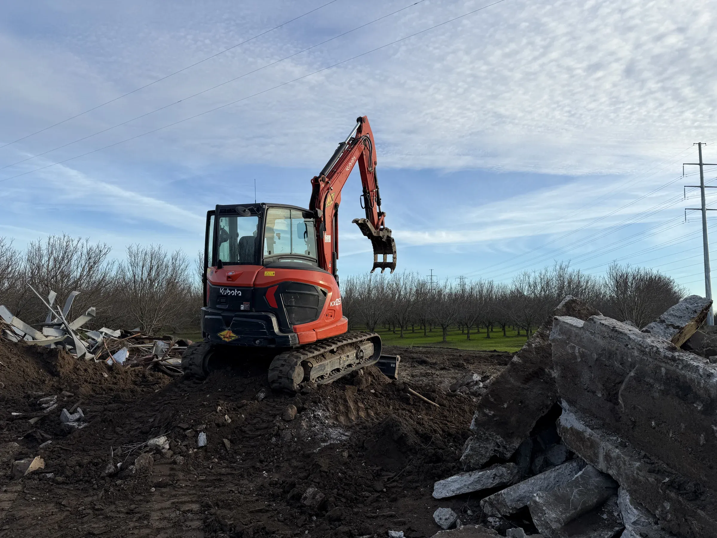 Demo Patrol Kubota excavator on demolition job site with concrete debris and Northern California orchard in the background