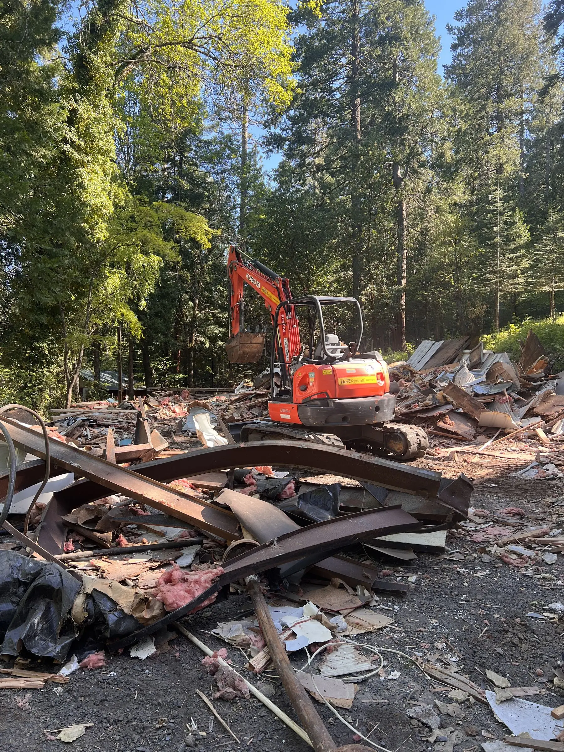 Excavator working through house demolition debris among tall pines